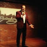 Shaun Eli, in a red jacket, on stage at Broadway Comedy Club