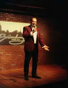 Shaun Eli, in a red jacket, on stage at Broadway Comedy Club