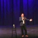 Corporate stand-up comedian Shaun Eli performing on a large stage in front of a purple curtain at Mamaroneck, New York's Emelin Theatre